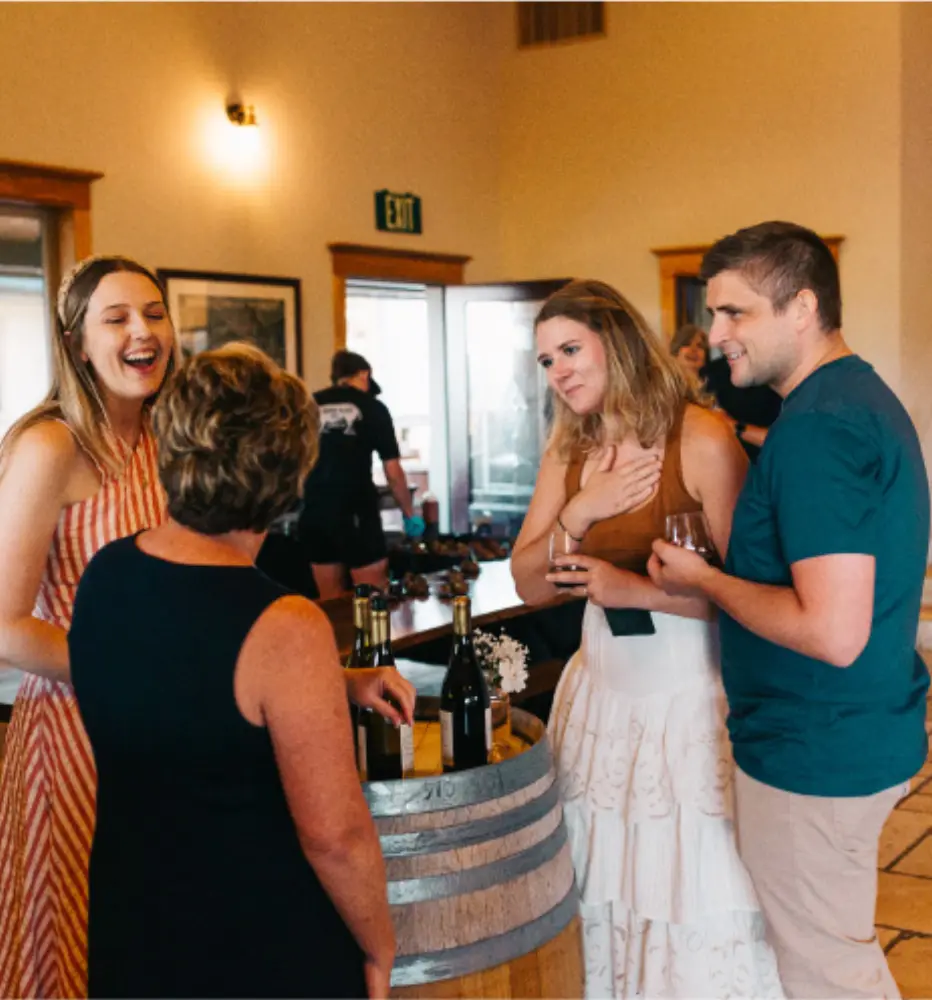 Guests laughing together with glasses of wine beside a barrel table in the Sweet Cheeks tasting room