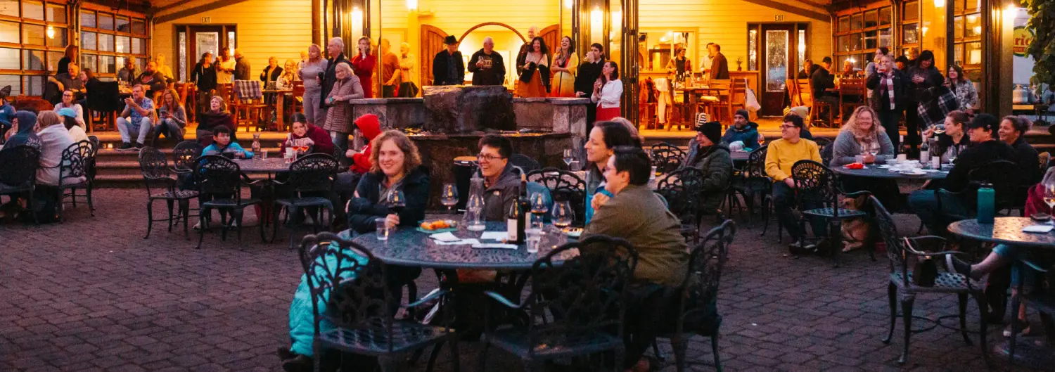 Guests seated at tables across the Sweet Cheeks patio at dusk, with the lit tasting room and a crowd gathered under the covered porch behind them